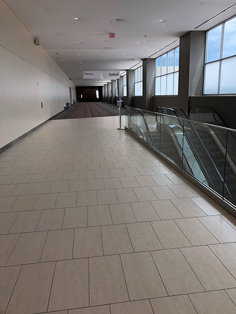 This photo shows a hallway. Extending approximately halfway down the hallway are rectangular beige tiles and then a gray and red striped carpet. On the left in this photo are escalators that lead down to the Meeting Room level.