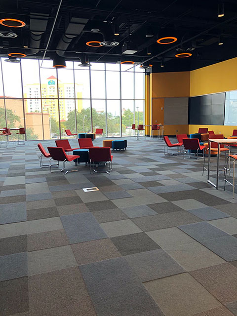 This is a photo of a large meeting room called the Cantilever Room. The carpet is made up of squares in varying shades of gray. The walls are yellow with floor-to-ceiling windows along one side. There are various types of seating in this space from low armchairs to high barstool-type chairs.