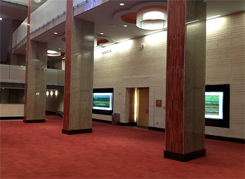 This photo shows the Lila Cockrell Theatre lobby. It has red carpet and rectangular granite columns. The entrance doors to the theater are interspersed through the lobby, are a light brown wood, and are located along a white tile wall.