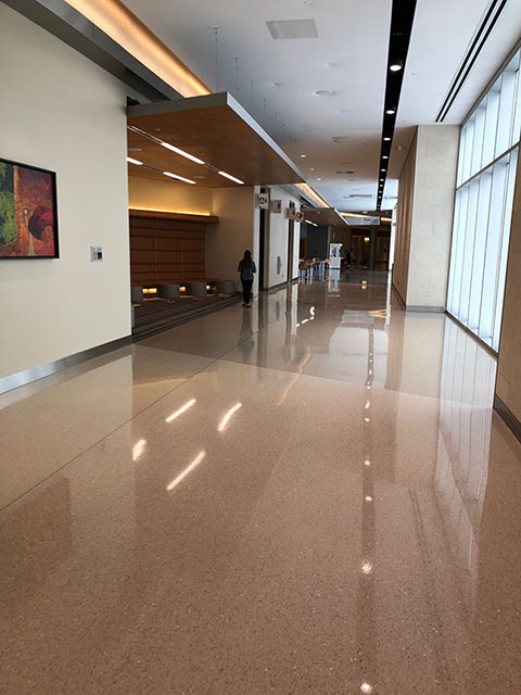 This photo shows the hallway between the Main Lobby and the West Lobby. The floor, like that in the Main Lobby, is made up of large beige speckled tiles. There are built-in seating areas on the left-hand side of the hallway that include benches, armchairs, and other mixed seating options. The nursing mothers’ room is also on the left of the hallway and has a sign posted above the door with a graphic indicating a mother and infant.