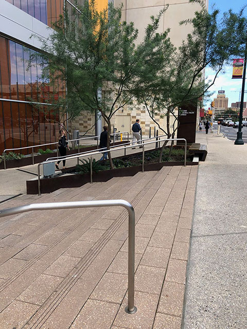 This photo shows a set of stairs from the Market Street sidewalk down to the plaza outside of the main entrance to the convention center. On either side of the stairs are silver handrails. Also in this photo is a ramp from the sidewalk down to the main entrance of the convention center. The ramp also has silver handrails on either side.