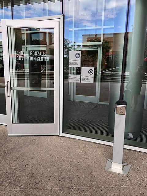 This photo shows a glass entry door to the convention center. There is a short pole to the right of the doorway with a wheelchair button.