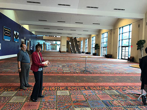 This photo shows a wide hallway with meeting rooms on the left side. The carpet is made up of individual squares, each with a unique pattern. There are also armchairs and couches on the left-hand side and doors leading out to a patio.