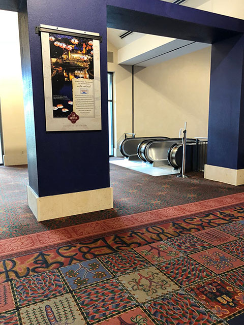 This photo shows the top of the escalators from the street level. The floor on the Meeting Room level is carpeted in a green, red, and yellow pattern. Down the hallway there is a patchwork pattern.