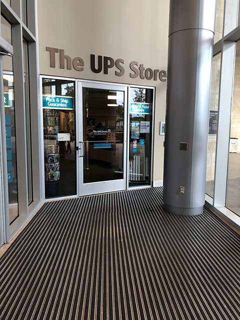 This photo shows the entrance to the UPS Store. The UPS Store has a glass entry door. The flooring before the entry is textured black-and-white stripe.