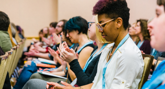 A row of people of sit in a full lecture hall, listening and taking notes